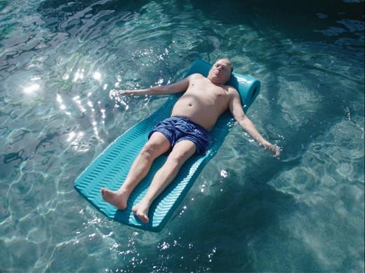 A masculine presenting person lying on a floating lounger in a pool of water with the sun shining on them.