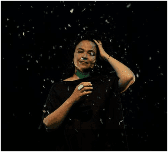 A woman in a black dress in front of a black background with what looks like snow falling in front of her.
