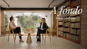 The image is a promotional still from a video titled "A Fondo: Sobre edadismo" (In Depth: About Ageism). It shows two women seated in chairs opposite each other, engaged in conversation, in what appears to be a library or study with extensive bookshelves. The setting is calm and intellectual, suggesting a serious discussion. The title card prominently displays the video's theme: ageism.
